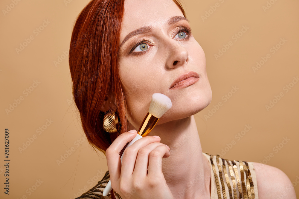redhead woman in gold earring applying face foundation with makeup brush on beige backdrop redhead woman in gold earring applying face foundation with makeup brush on beige backdrop