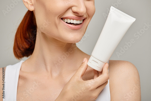 cropped view of cheerful woman with red hair holding tube with body lotion on grey background