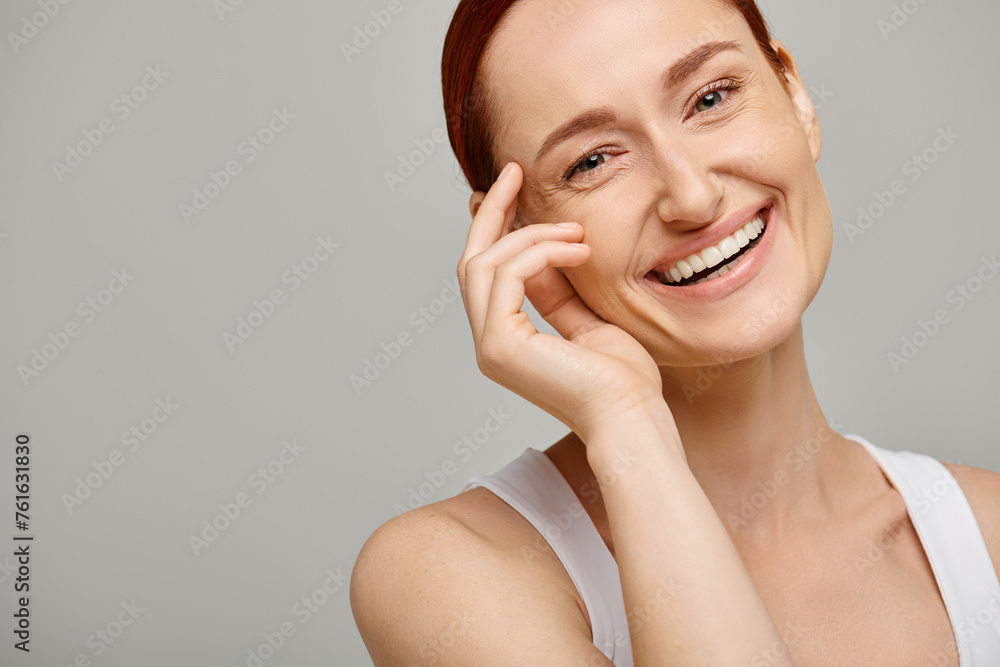 cheerful and redhead woman in white tank top looking at camera on grey background, healthy smile cheerful and redhead woman in white tank top looking at camera on grey background, healthy smile