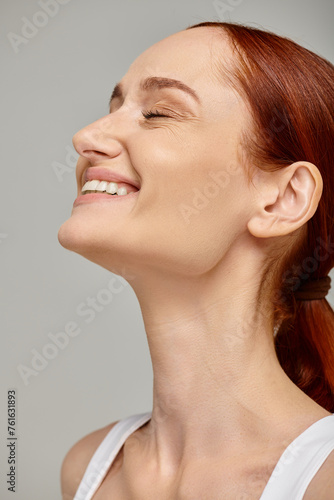 happy redhead woman with closed eyes exuding joyful and healthy smile on grey background © LIGHTFIELD STUDIOS