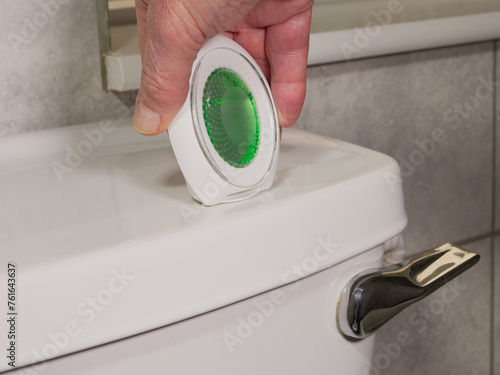 Closeup of a man’s hand placing a scented oil air freshener on the water cistern lid of a bathroom toilet.
