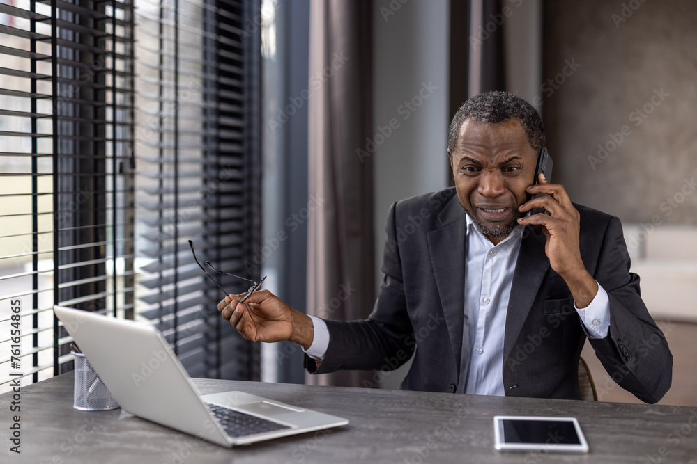 Furious diverse male in formalwear holding eyeglasses while having unpleasant phone conversation. Angry distant worker hearing bad breaking news while sitting by desktop with portable computer Furious diverse male in formalwear holding eyeglasses while having unpleasant phone conversation. Angry distant worker hearing bad breaking news while sitting by desktop with portable computer