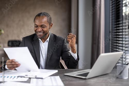 Happy diverse male raising hand with clenched fist while looking at document in hand by desktop with pc. Rejoiced manager reading information with approving result of salary increasing request