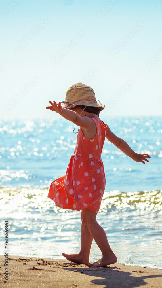 Happy little girl with raised arms walks along the beach in summer back to the camera. Baby in an orange dress with polka dots and a straw hat. Vertical banner Happy little girl with raised arms walks along the beach in summer back to the camera. Baby in an orange dress with polka dots and a straw hat. Vertical banner