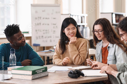 Portrait of smiling Asian girl sitting at table with diverse group of students and enjoying schoolwork in college library