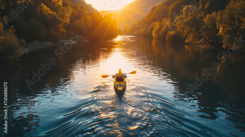 A person kayaking down a tranquil river, basking in the golden glow of the sunset with forested hills in the background. © khonkangrua