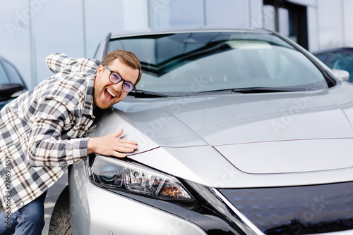 Young bearded man embracing his new car at the dealership smiling joyfully