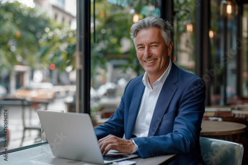 A high-resolution photo captures a middle-aged businessman, dressed in a blue suit and tie, smiling as he types on his laptop at a modern cafe with a cityscape view © gabriele
