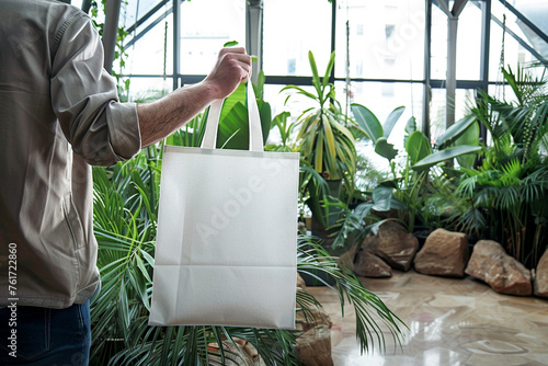 A man holding a blank white tote, reusable shopping bag in a dynamic, modern atrium filled with plants, underlining the integration of nature into architectural design, photo