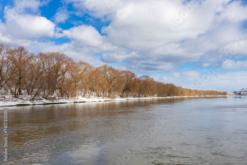 Trees along the river, clouds reflected in the water, spring on the river, salty weather, high water, view from the middle of the river to the shore. Blue sky with clouds