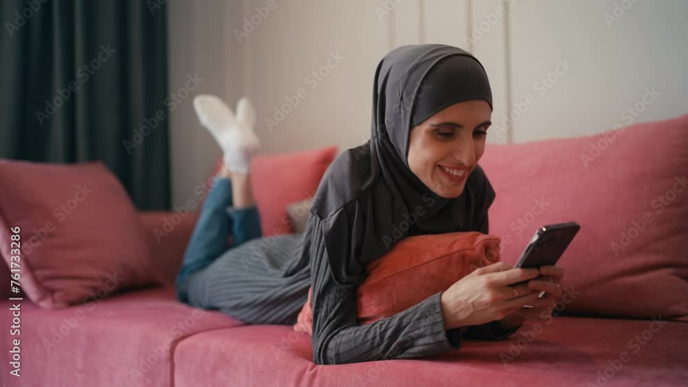 Pretty young female in hijab lying on couch, chatting in messenger app on gadget Pretty young female in hijab lying on couch, chatting in messenger app on gadget