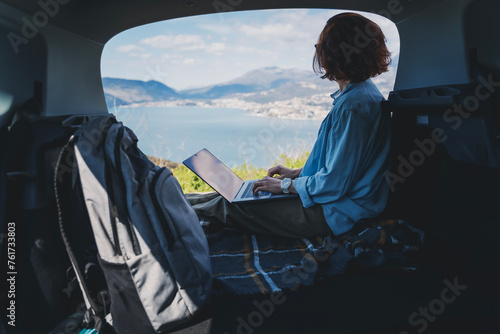 Young woman sitting in the open trunk of a car overlooking the sea and using a laptop, freelancer, remote work and summer vacation auto travel © olezzo
