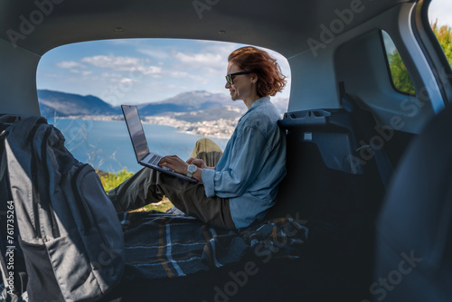 Young woman sitting in the open trunk of a car overlooking the sea and using a laptop, freelancer, remote work and summer vacation auto travel © olezzo