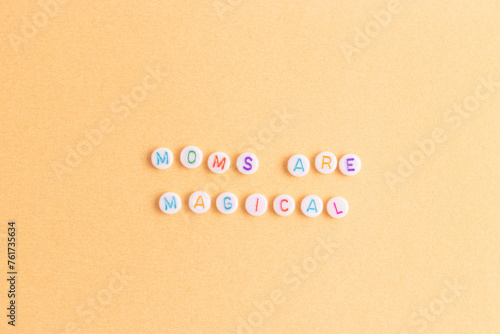 Moms are magical. Quote made of white round beads with multicolored letters on a gold colored background.