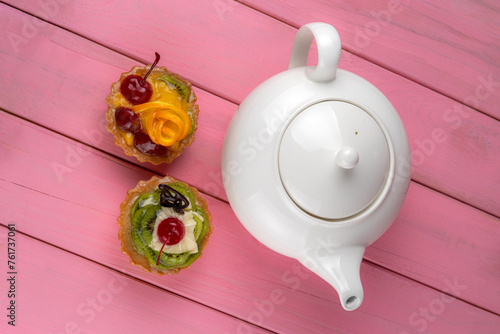 Colorful fruit cakes made with kiwi, orange, candied cherry, cream and chocolate on a pink background with a teapot.
