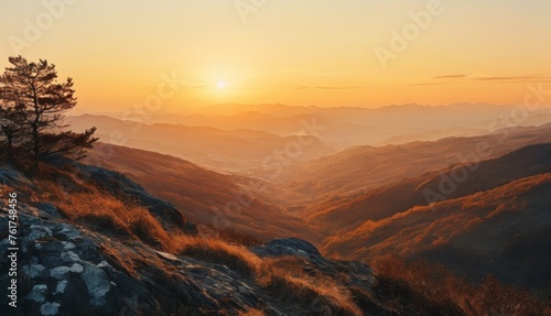  the sun setting over a mountain range with a lone tree on the top of a hill in the foreground. © Jevjenijs