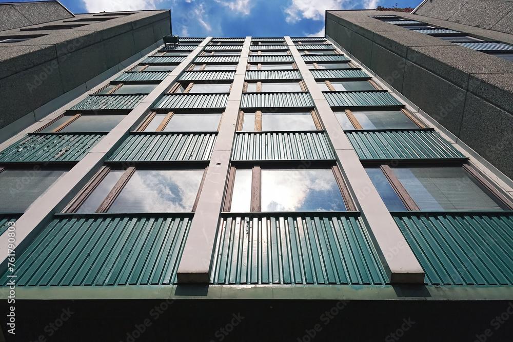 Fototapeta premium Wall of a multi-storey building with reflection of the sky and sunlight in one of the windows exterior. High building in the city, view from below. Office building with sky. 