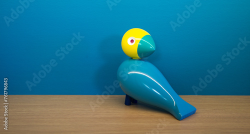 A colorful toy bird made of wood sitting on a shelf in front of a blue wall. The head of the bird is yellow.  photo