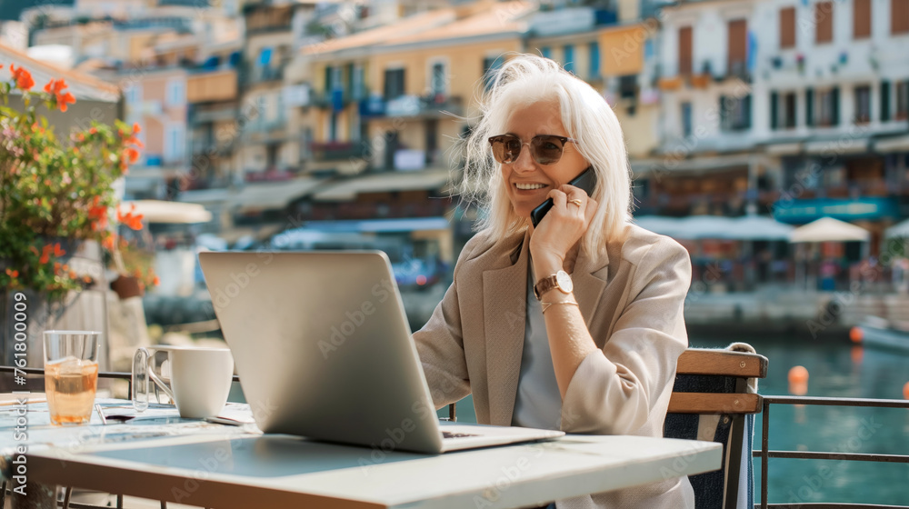 Blonde elderly woman in a cafe on the waterfront near the marina, working online, talking on a smartphone with business partners during lunch Blonde elderly woman in a cafe on the waterfront near the marina, working online, talking on a smartphone with business partners during lunch