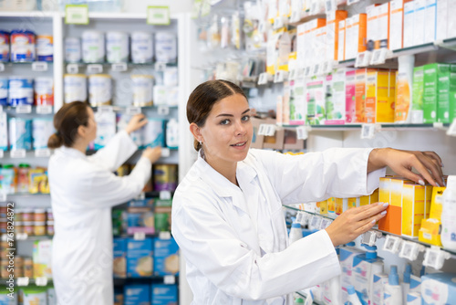 Young woman pharmacist in uniform arranges products on shelves in pharmacy