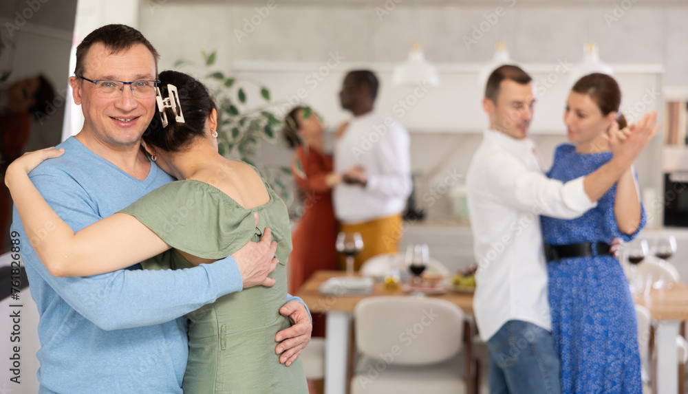 Fototapeta premium Smiling middle-aged man in blue jumper and glasses enjoying slow dance with brunette in olive dress at relaxed friendly home gathering