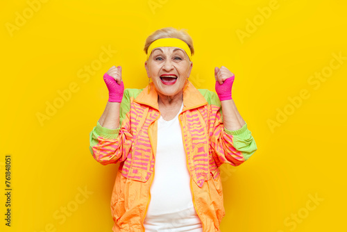 crazy funny old granny in sports colorful clothes wins and celebrates success on a yellow isolated background, elderly woman in youth hipster clothes rejoices in victory and luck and screams