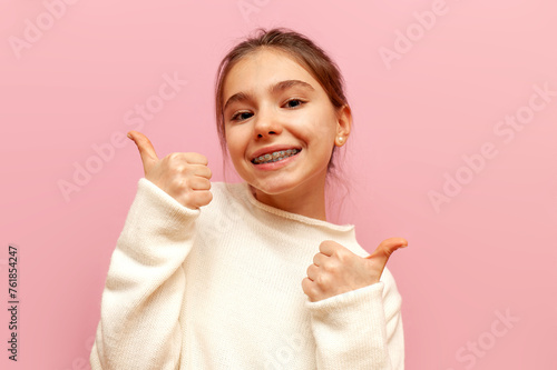 cheerful teenage girl with braces smiles and shows likes with her hands on a pink isolated background, child shows thumbs up and recommends