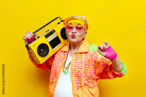 funny crazy granny in hipster clothes listening to music on a tape recorder and showing a kiss on a yellow isolated background, elderly cool woman dancing at a party and having fun