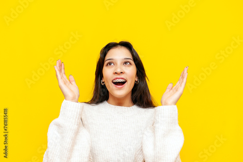 young shocked asian woman with braces wonders and holds empty hands over yellow isolated background, korean girl raises her hands up in amazement