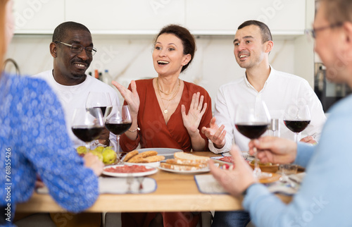 Happy middle-aged men and women sitting at table with a glass of wine while having friendly conversation at home