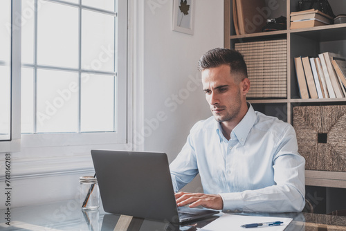Happy young caucasian millennial businessman working at home at desk with laptop or computer having fun. Young male person in video conference taking care of the business or market..