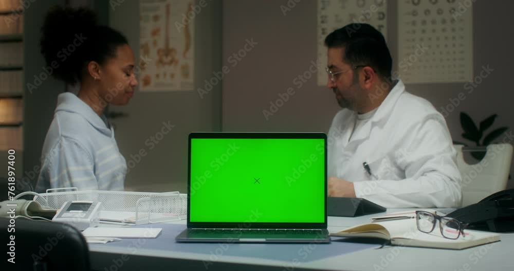 A laptop with a green screen is on the table in the medical office. The doctor measures the temperature of a female patient A laptop with a green screen is on the table in the medical office. The doctor measures the temperature of a female patient