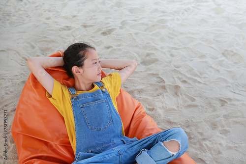 Portrait of Asian young girl in dungarees jean looking beside and relax on orange sofa bed beach on sand at summer holiday.