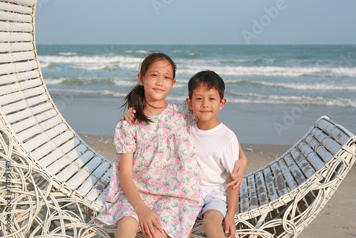 Portrait of happy Asian boy and girl sitting and hugging each other on white bamboo wooden sofa bed and looking camera on the beach at summer holiday.