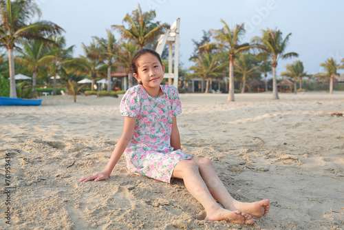 Portrait of happy Asian young girl child relax on beach sand and looking camera at summer holiday.