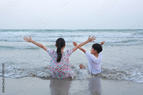 Back view of Asian young girl child and little brother sitting and raised hands up on tropical sand beach at sunset. Adorable sister and brother having fun in summer holiday.