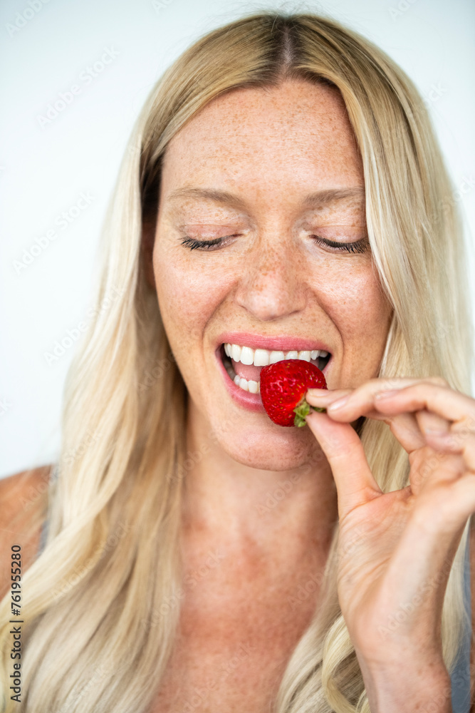 Blonde woman with freckles smiling biting on a strawberry Blonde woman with freckles smiling biting on a strawberry