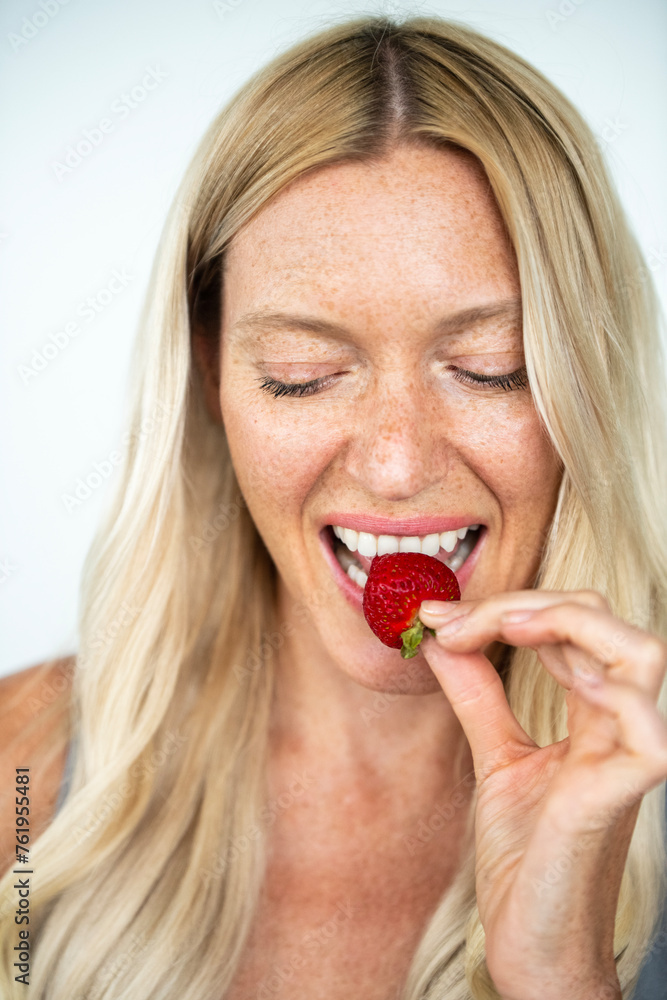 Blonde woman with freckles smiling biting on a strawberry Blonde woman with freckles smiling biting on a strawberry