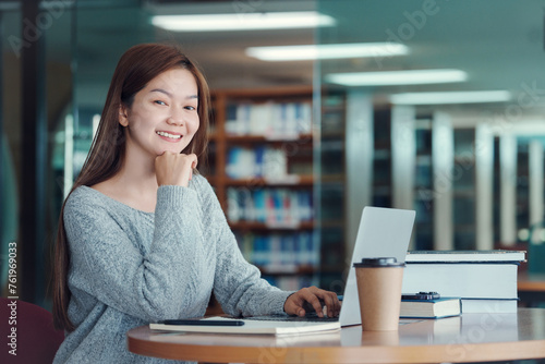 Happy young girl student studying at the college library, sitting at the desk, using laptop computer.