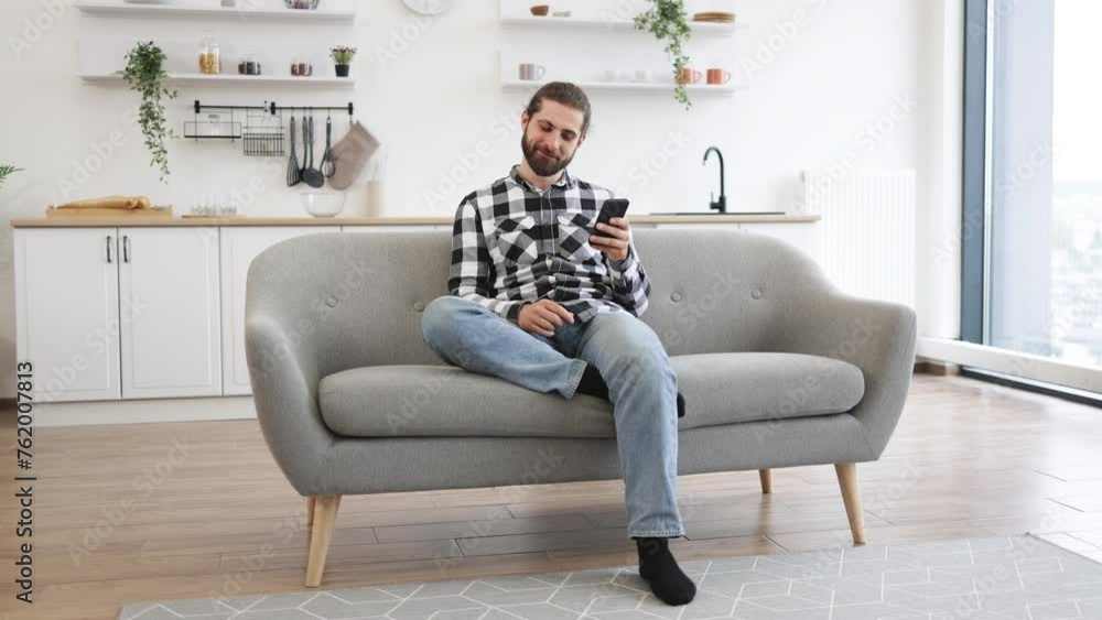Caucasian male in checkered shirt holding smartphone while staying in modern workplace at home. Efficient freelance worker texting message to colleague while performing project for employer indoors. Caucasian male in checkered shirt holding smartphone while staying in modern workplace at home. Efficient freelance worker texting message to colleague while performing project for employer indoors.