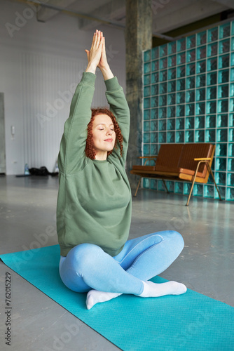 Vertical image of pretty relaxed female in sports clothes practicing yoga indoor at gym, sitting on blue mat in lotus position with closed eyes and raised hands keeping palms folded, breathing deeply