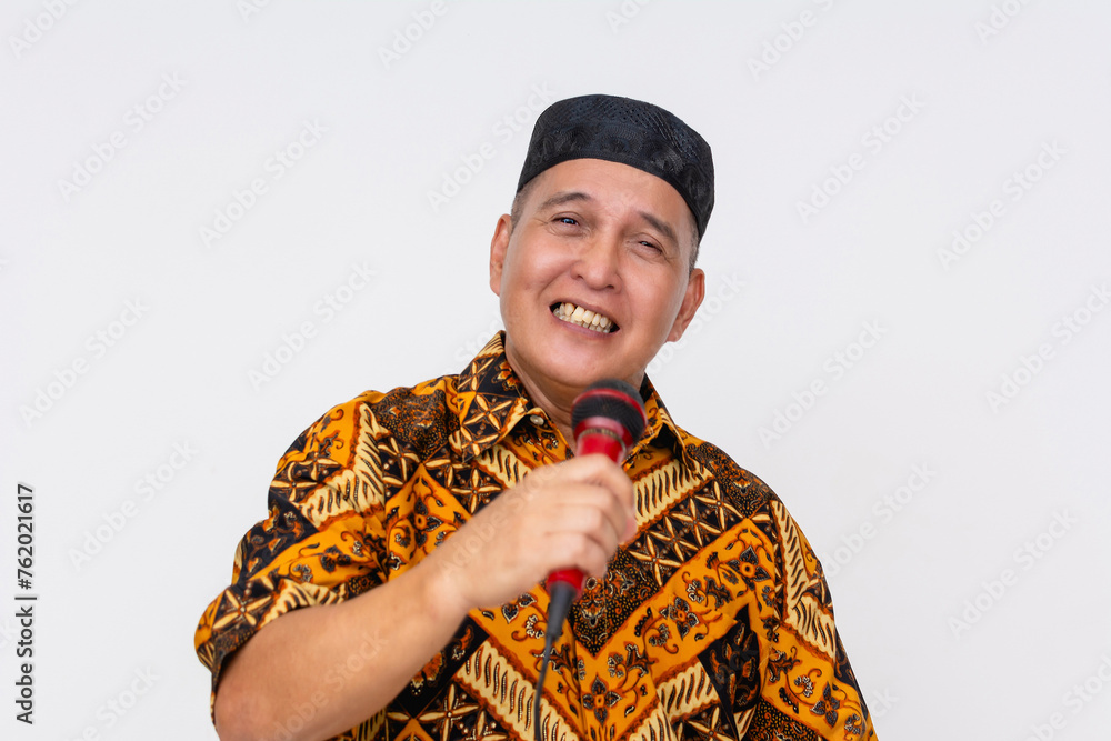 Joyful Indonesian man wearing traditional batik shirt and kopiah hat singing into a microphone on a white background. Joyful Indonesian man wearing traditional batik shirt and kopiah hat singing into a microphone on a white background.