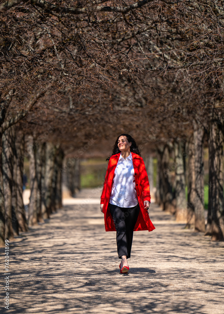 stylish, confident woman outdoors. A model with dark hair, dressed in a red coat, white blouse and red high-heeled shoes, cheerfully flies along an alley in the park. Spring fashion trend
