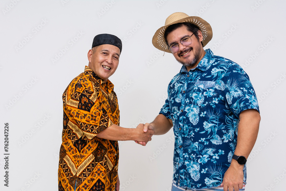 A smiling Indonesian man in traditional batik attire and kopiah hat warmly greets a tourist in a vibrant hawaiian shirt, handshake gesture captured on white background. A smiling Indonesian man in traditional batik attire and kopiah hat warmly greets a tourist in a vibrant hawaiian shirt, handshake gesture captured on white background.