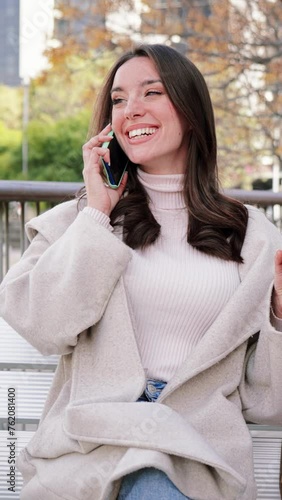 Vertical. One brunette lady doing a call with a cell phone sitting on a bench in a city street after shopping. Isolated young woman. Smiling and talking with a smar tphone. Communication. Slow motion photo