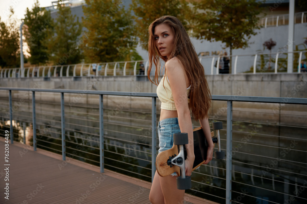 Sensual slim young woman walking with longboard over summer cityscape