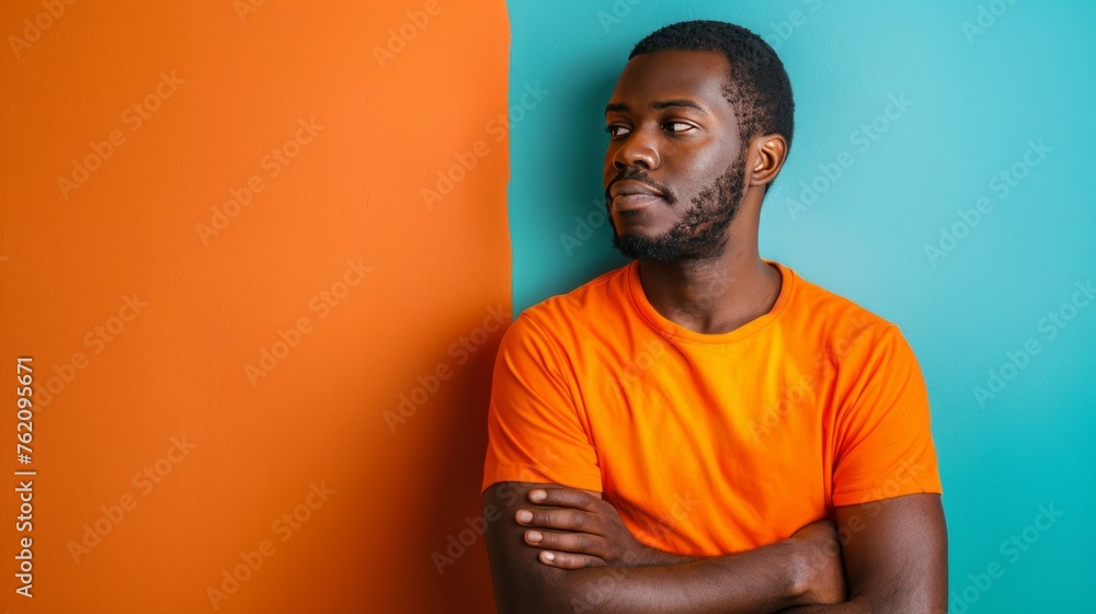 an African American man in orange t-shirt is standing against an orange and blue wall. He is pensive, thoughtful, worried, looking in the distance an African American man in orange t-shirt is standing against an orange and blue wall. He is pensive, thoughtful, worried, looking in the distance