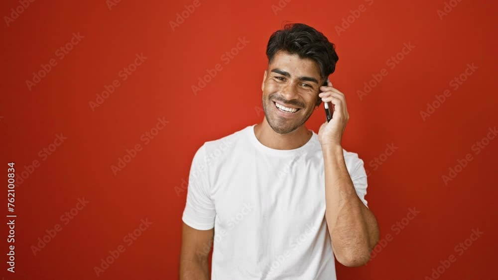 A handsome young hispanic man smiles while talking on the phone against a red wall. A handsome young hispanic man smiles while talking on the phone against a red wall.