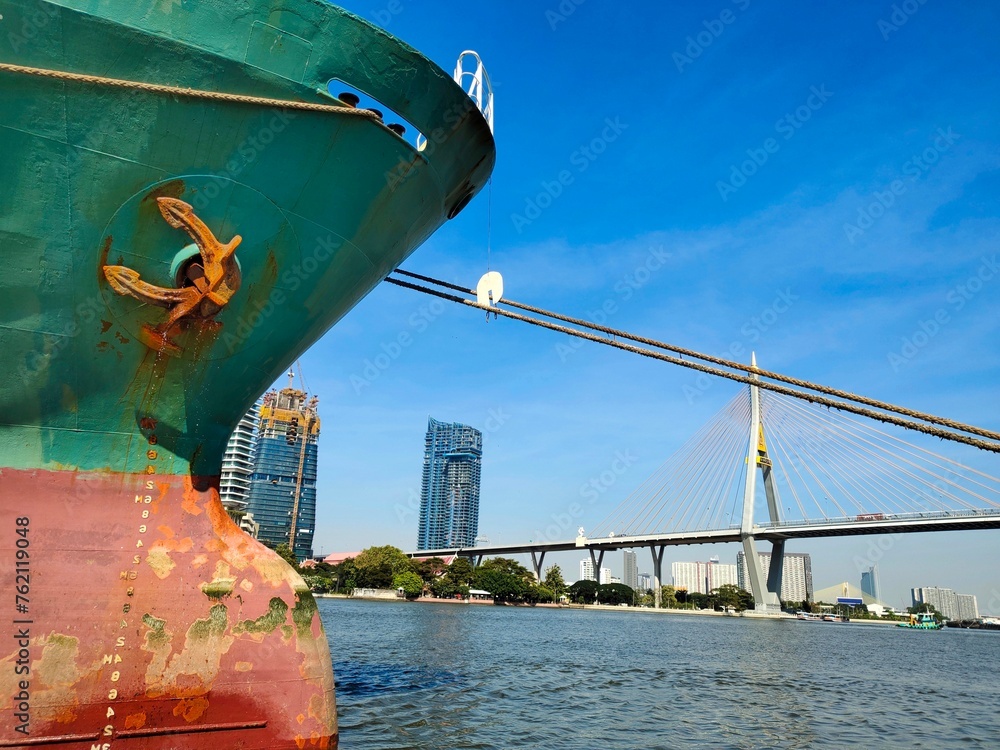Naklejka premium ship in the port, Anchor on large cargo ship's anchor being pulled. Green and red ship, While docked at the pier by large ropes on the river, on the building background and transport concept