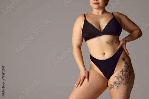 Cropped portrait of female plus-size model posing in dark bikini, underwear against grey studio background. Copy space. Concept of natural beauty, femininity, body positivity, dieting, fitness. © Lustre